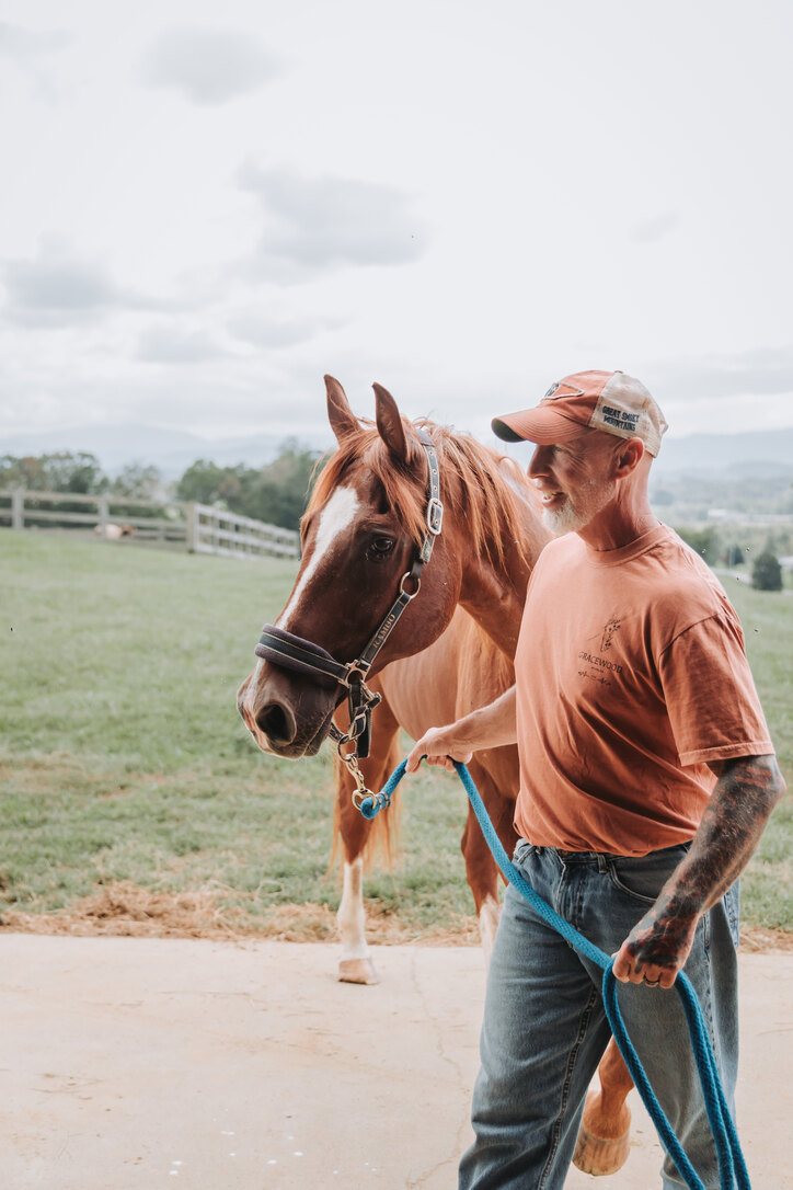 Robbie in his Gracewood tee leading a horse in from the pasture, the mountains behind